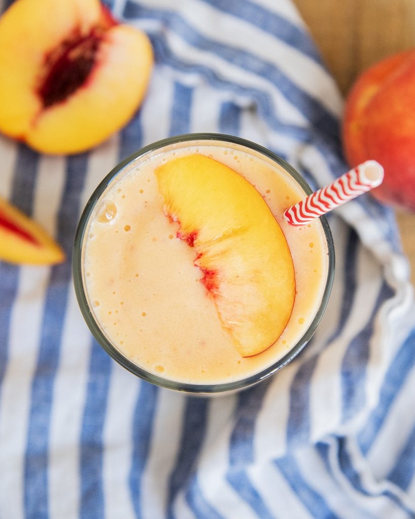 An overhead photo of a glass of a peach smoothie with a peach slice on top, and a red straw in it.