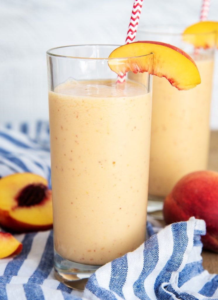 Two tall glasses of peach smoothie with a peach slice on top of the edge of the glass.