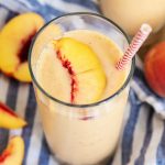 A glass full of peach smoothie with a slice of a peach on top.
