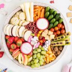 An overhead photo of a round snack board filled with fruits and veggies, sandwiches, cheese, and cookies.