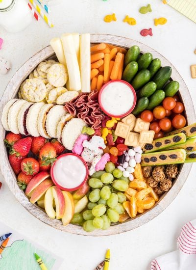 An overhead photo of a round snack board filled with fruits and veggies, sandwiches, cheese, and cookies.
