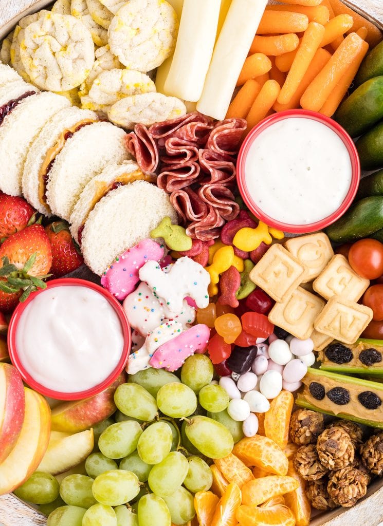 A close up of a snack charcuterie board with two bowls of dips and fruit and veggies, and crackers.