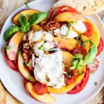 A peach and tomato burrata salad on a plate, with the burrata cut into, and topped with balsamic glaze.