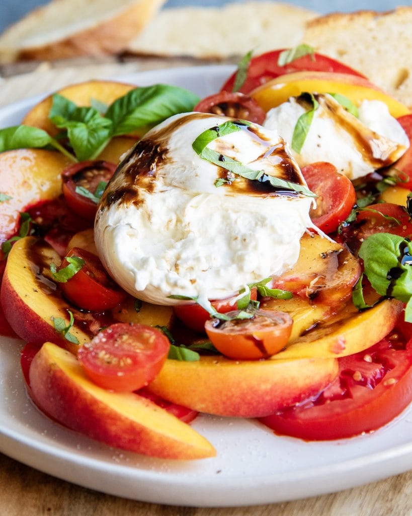 A plate of peach burrata salad with slices of tomatoes, and topped with balsamic glaze and fresh basil.