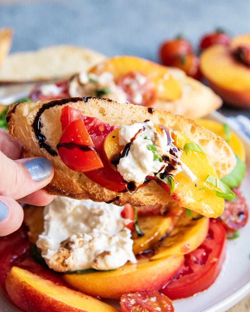 A piece of toasted baguette with burrata cheese, and sliced peaches, and tomatoes on top.