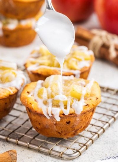 Vanilla icing being drizzled by a spoon onto a cinnamon roll apple pie cup.