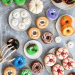 An overhead photo of 9 varieties of Halloween donuts, there are monster donuts, vampire donuts, eyeball donuts, spider web donuts, pumpkin donuts, and more.
