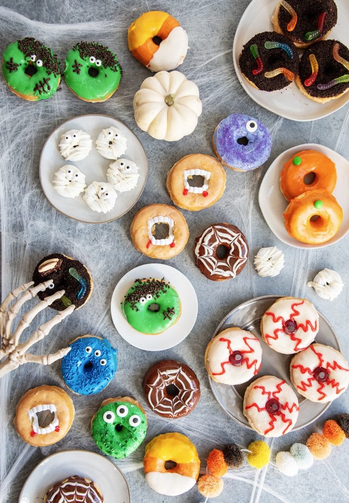 An overhead photo of 9 varieties of Halloween donuts, there are monster donuts, vampire donuts, eyeball donuts, spider web donuts, pumpkin donuts, and more.
