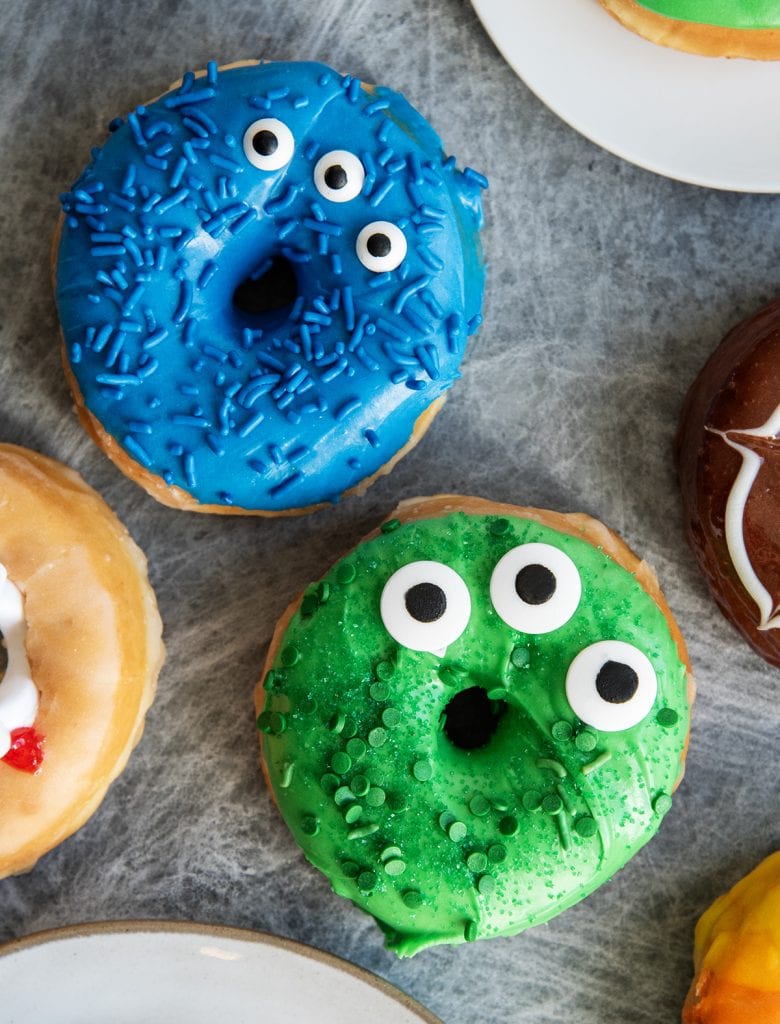Two monster donuts, one is blue, one is green with three eyes, and sprinkles on top.