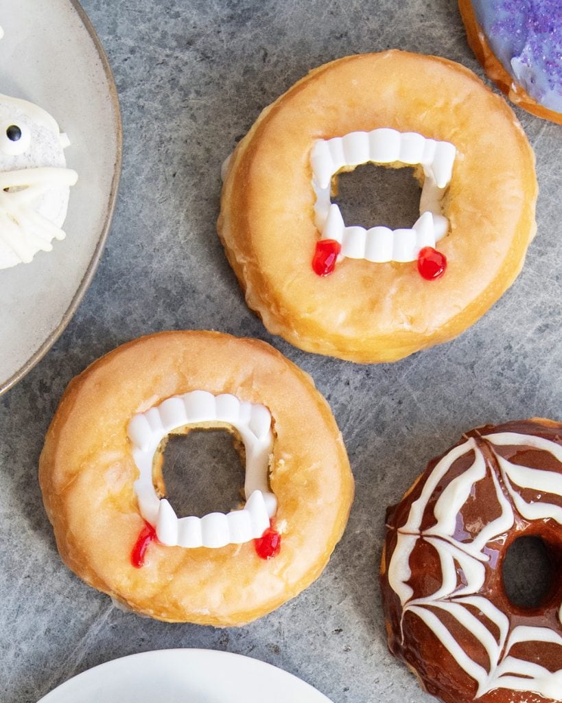 Two glazed donuts decorated to look like vampire mouths with plastic fangs and red gel looking like blood.