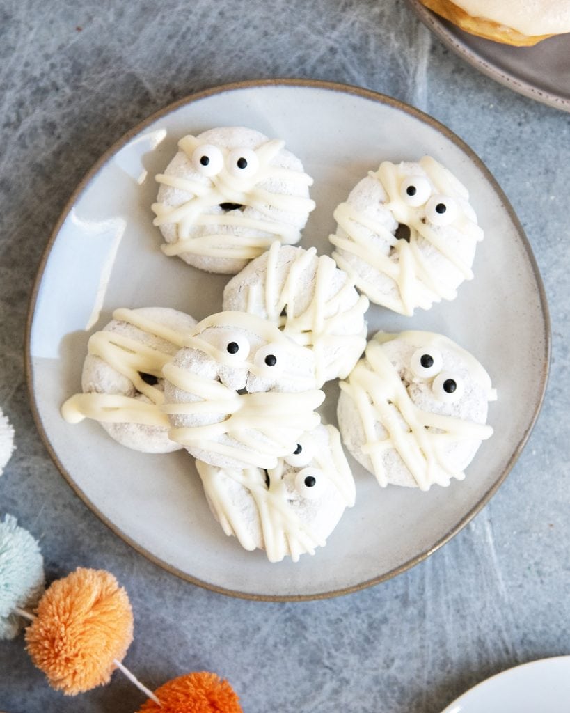 A plate of mini powdered drizzled with white chocolate, and topped with candy eyes to look like mummies.