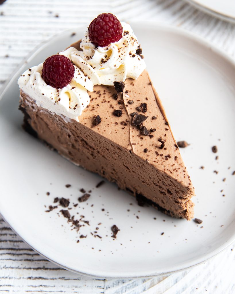 A slice of chocolate cheesecake on a plate, topped with whipped cream and two raspberries.