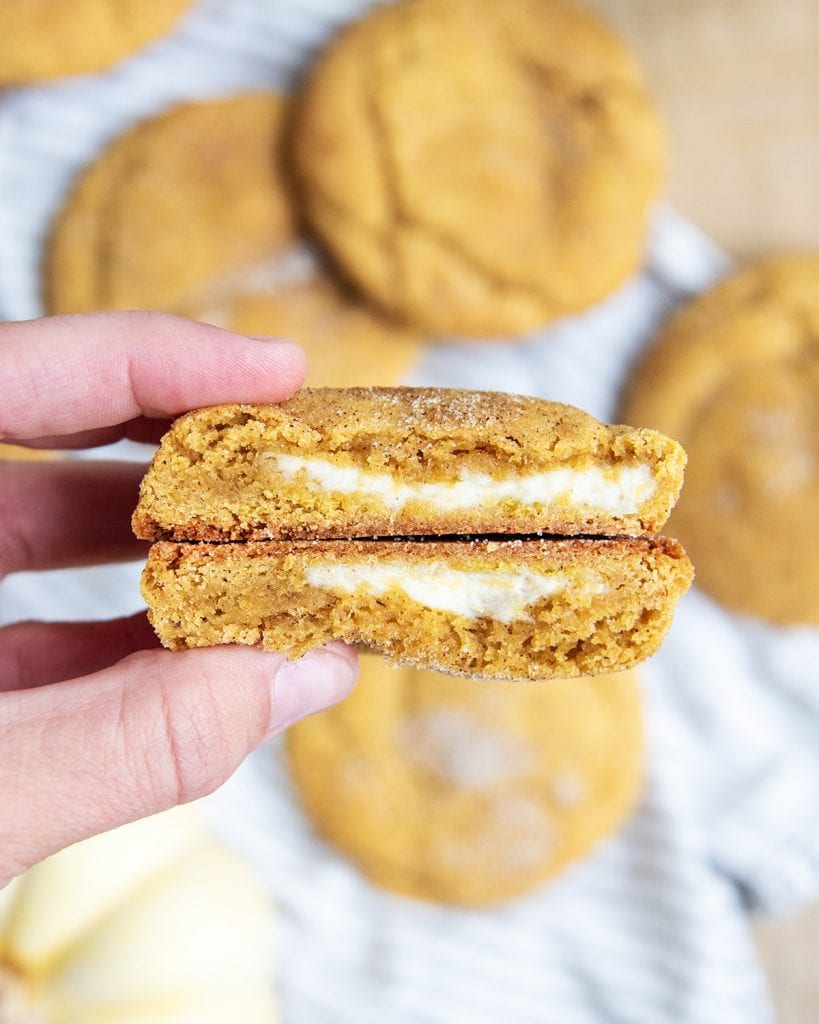 A hand holding two halves of a pumpkin cheesecake cookie, showing the cheesecake filling in the middle.