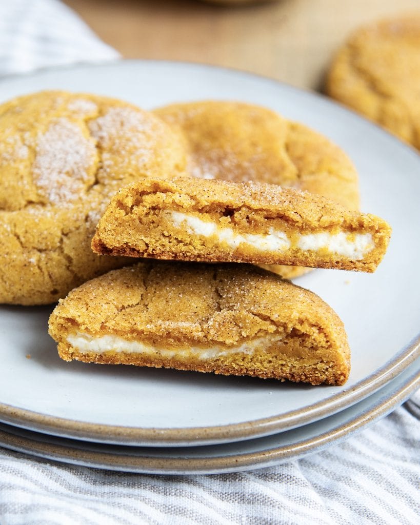 A plate with two halves of a pumpkin cream cheese cookie with the cheesecake filling showing in the middle of the pumpkin cookie.