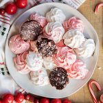 A plate of pink and white peppermint meringue cookies, some are flipped over, showing they are dipped in chocolate and coated in candy cane pieces.