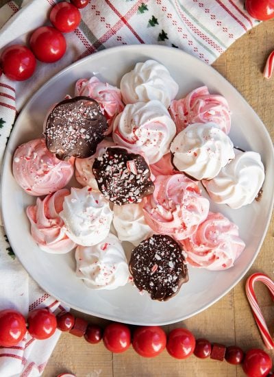 A plate of pink and white peppermint meringue cookies, some are flipped over, showing they are dipped in chocolate and coated in candy cane pieces.