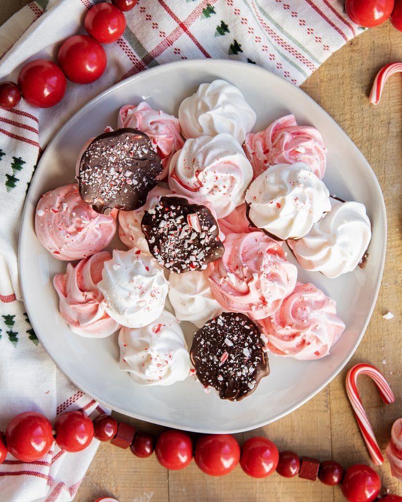 A plate of pink and white peppermint meringue cookies, some are flipped over, showing they are dipped in chocolate and coated in candy cane pieces.