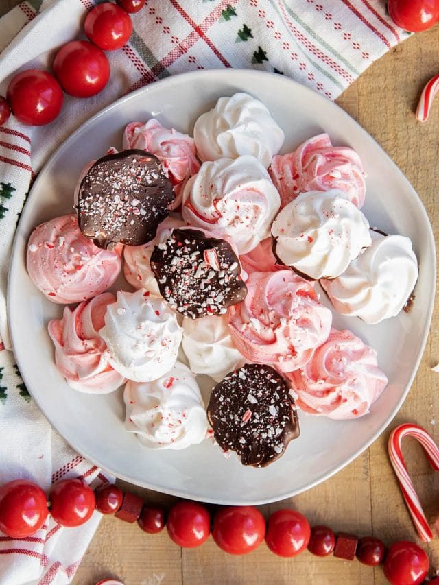 A plate of pink and white peppermint meringue cookies, some are flipped over, showing they are dipped in chocolate and coated in candy cane pieces.
