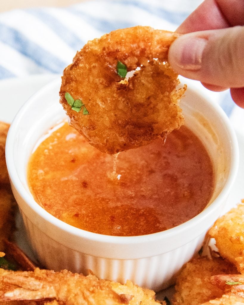 A hand dipping a coconut shrimp into a bowl of thai chili sauce.