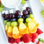 A tray of rainbow colored fruit kabobs with a marshmallow on the end, and a bowl of white colored fruit dip.