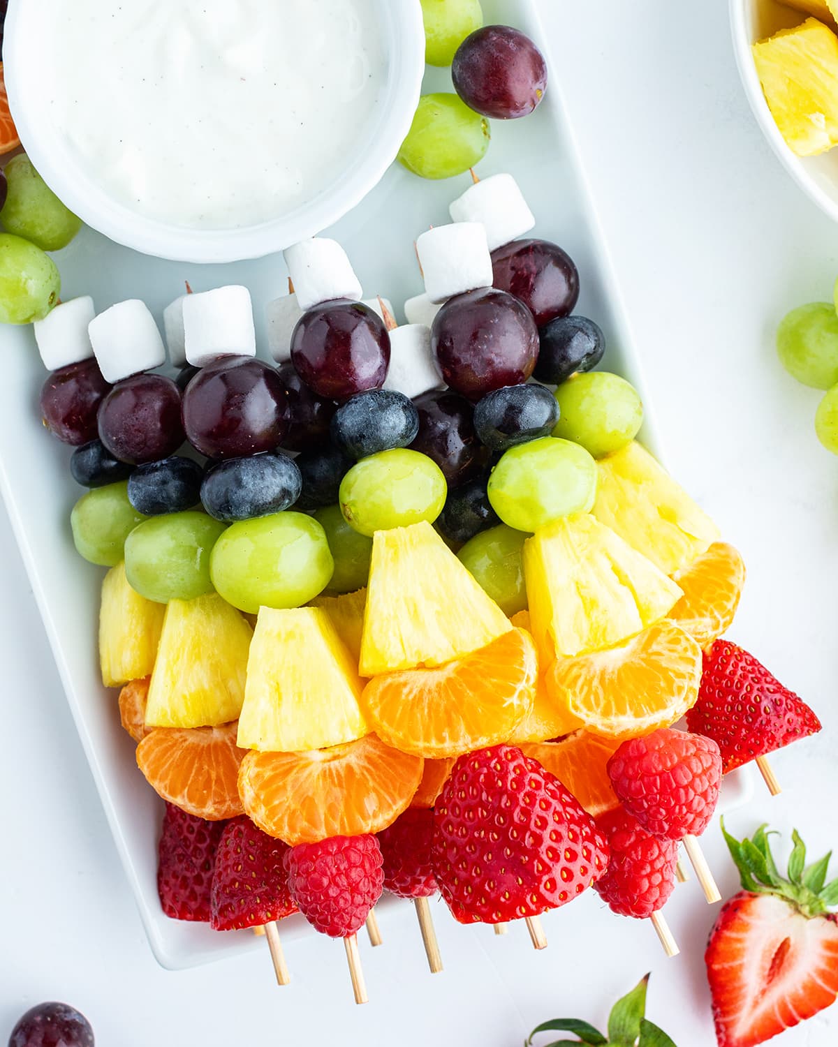 A tray of rainbow colored fruit kabobs with a marshmallow on the end, and a bowl of white colored fruit dip.