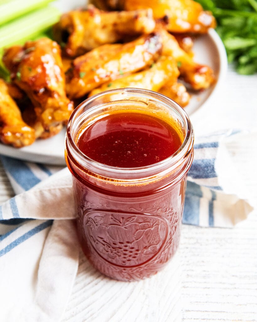 A pint glass jar of homemade buffalo wing sauce, with a plate of wings behind them.