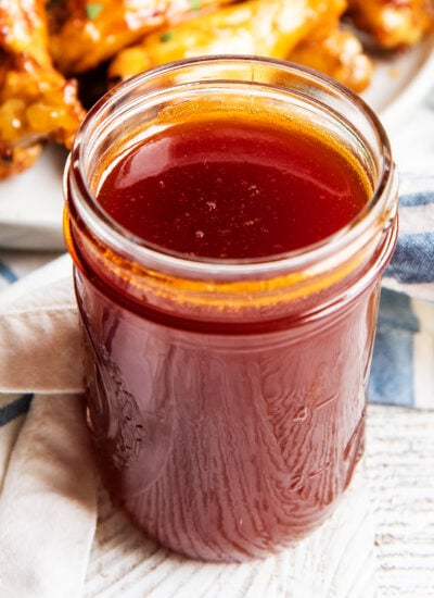 A glass jar full of homemade honey buffalo wing sauce, with a plate of wings behind it.