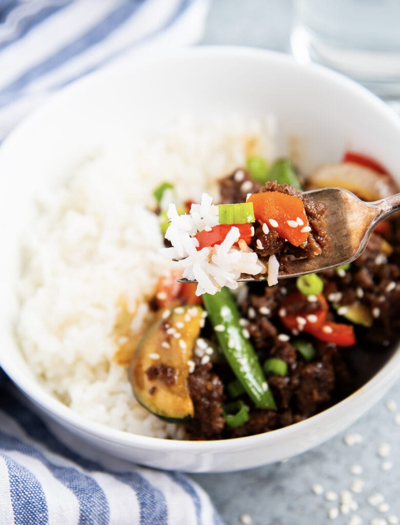 A bowl of rice with ground beef and vegetable stir fry, and a bite on a fork in front.