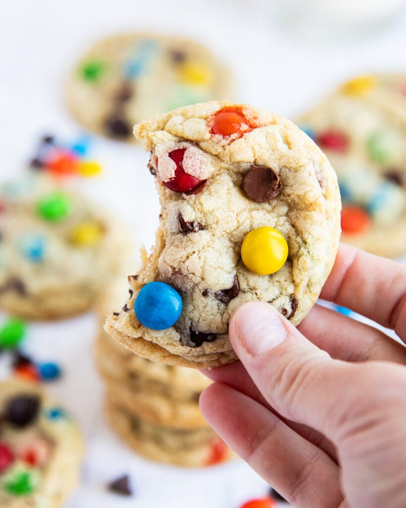 A hand holding a M&M and chocolate chip cookie with a bite out of it.