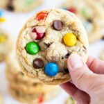 A hand holding a M&M cookie topped with a green, blue, yellow, and orange m&m and chocolate chips.