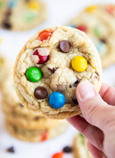 A hand holding a M&M cookie topped with a green, blue, yellow, and orange m&m and chocolate chips.