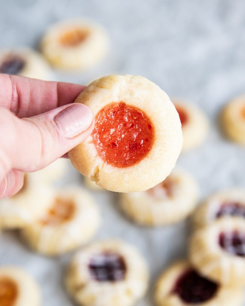 A hand holding a small thumbprint cookie filled with strawberry jam.