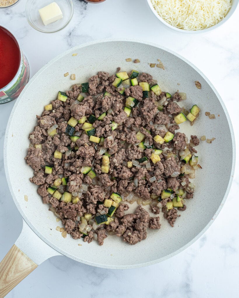 A white saute pan with browned ground beef and zucchini.