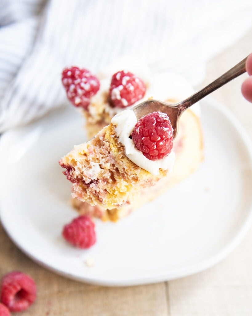 A bite of cake on a plate topped with cream cheese frosting, and a fresh raspberry on a plate.
