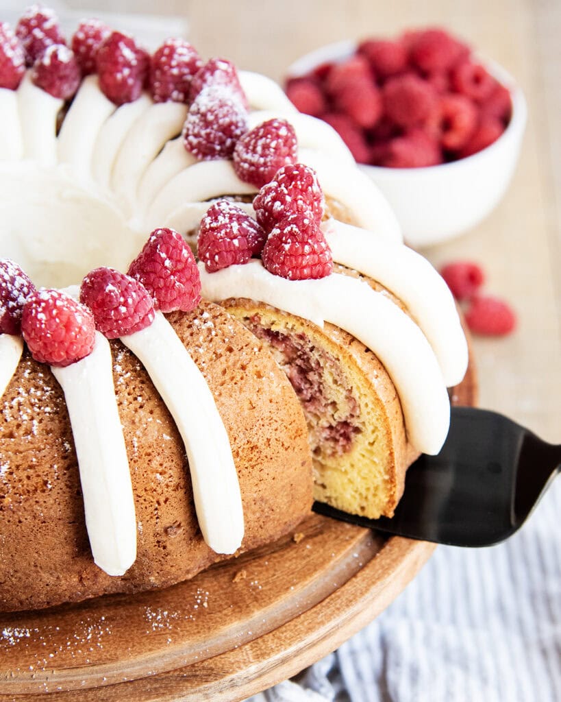 A raspberry bundt cake topped with cream cheese frosting, with a piece being removed with a metal spatula.