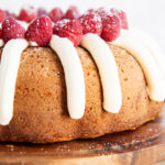 A bundt cake on a wooden cake stand topped with piped cream cheese frosting, and fresh raspberries.