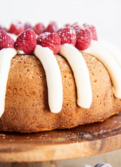 A bundt cake on a wooden cake stand topped with piped cream cheese frosting, and fresh raspberries.