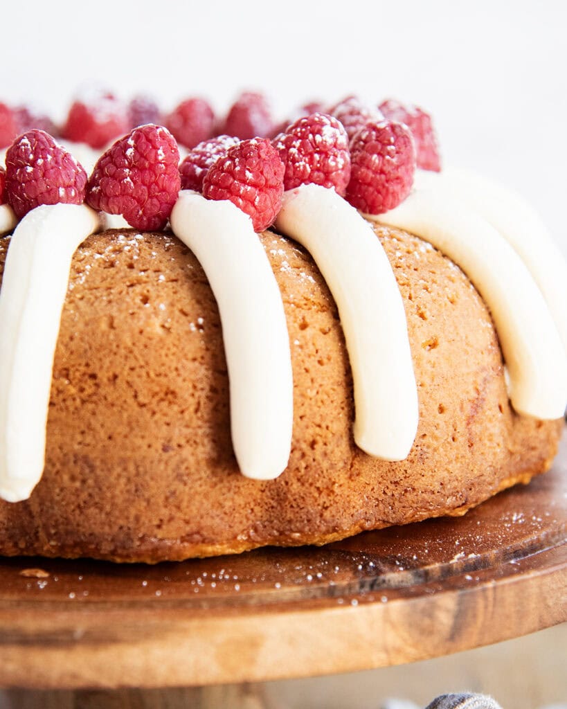 A bundt cake on a wooden cake stand topped with piped cream cheese frosting, and fresh raspberries.