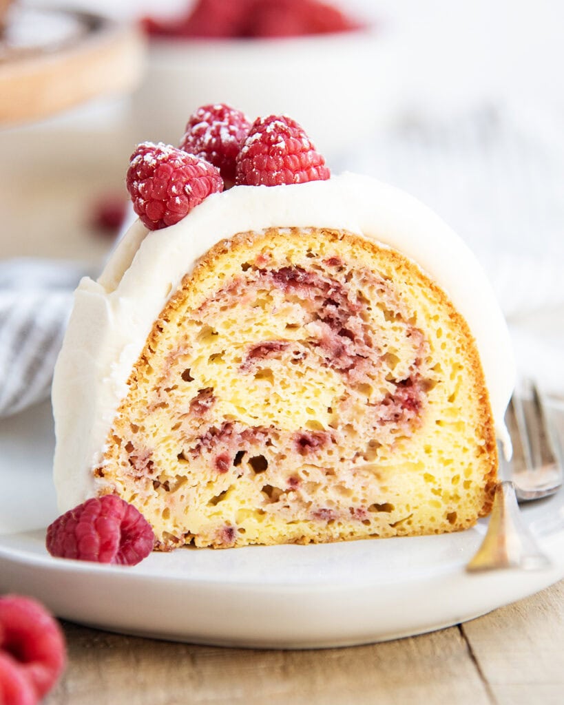 A close up of a piece of a raspberry white chocolate bundt cake on a plate topped with cream cheese frosting and three fresh raspberries.