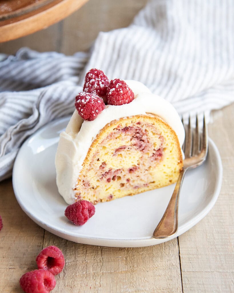A piece of a raspberry bundt cake on a plate topped with cream cheese frosting and three fresh raspberries.