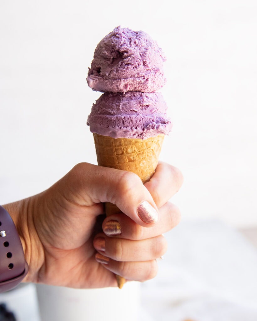 A hand holding a cone of blueberry ice cream, with 2 scoops.