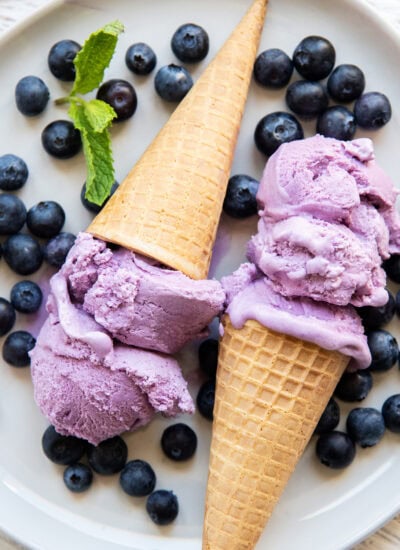 Two ice cream cones full of blueberry ice cream on a plate with fresh blueberries on the plate.