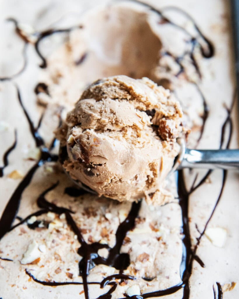 A scoop of mocha ice cream on top of a container of ice cream with fudge swirls around.