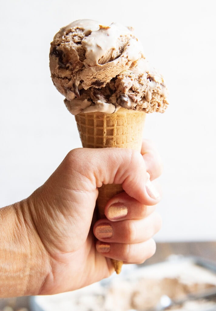 A hand holding an ice cream cone topped with a chocolatey fudge swirled ice cream.