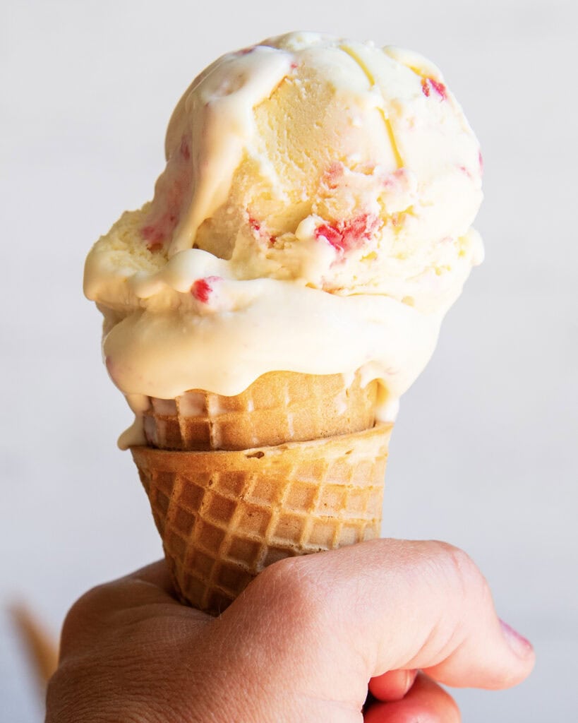 A hand holding an ice cream cone topped with white chocolate raspberry ice cream.