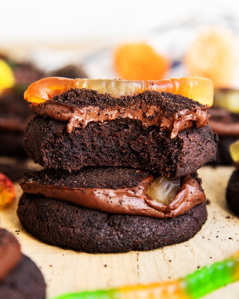 A thick frosted chocolate cookie with a bite out of it, and a gummy worm on top, with another cookie stacked underneath.