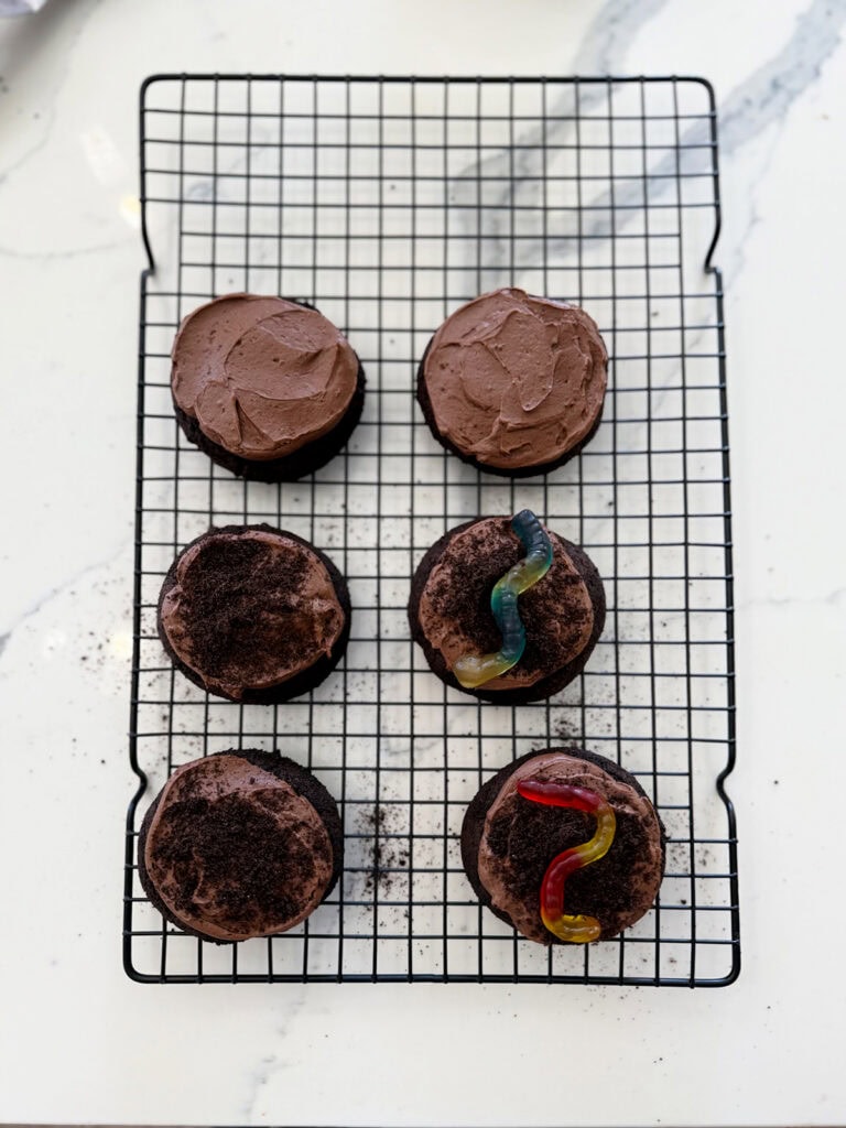 Dirt cake cookies topped with Oreo crumbs and gummy worms on a wire rack.