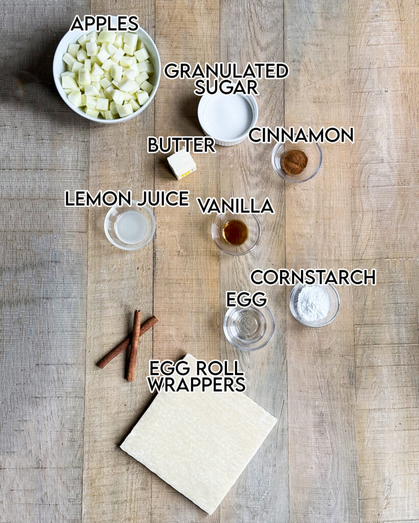 Small bowls of the ingredients needed to make apple pie egg rolls on a wooden table.