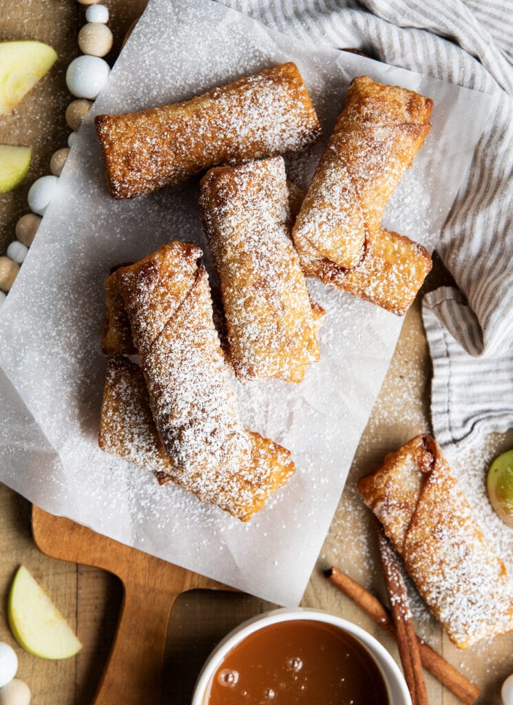 An above view of apple pie egg rolls on parchment paper next to a bowl of caramel dip.
