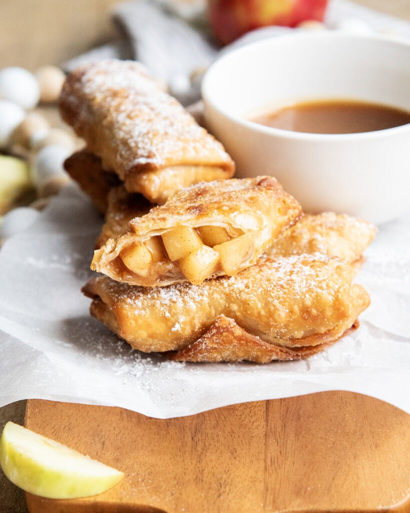 Apple pie egg rolls dusted in powdered sugar and one is cut in half showing the apple pie filling.